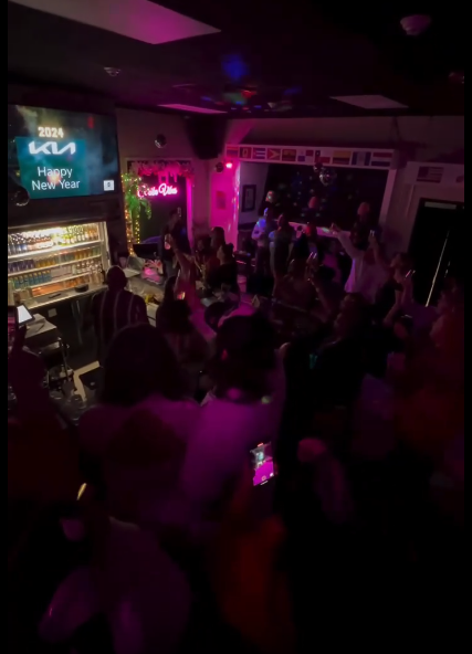 Packed Caribe Happy New Year 2024 party with neon lights, the bar crowd cheering, and the big screen